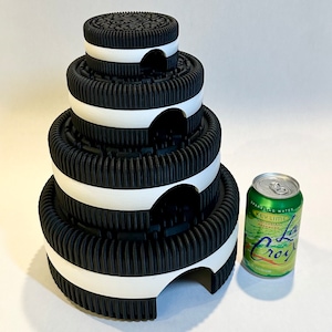 May include: A stack of four black and white Oreo cookies, each with a white rim, stacked on top of each other. The cookies are arranged in a pyramid shape. The cookies are on a white surface. A can of sparkling water is in the background.