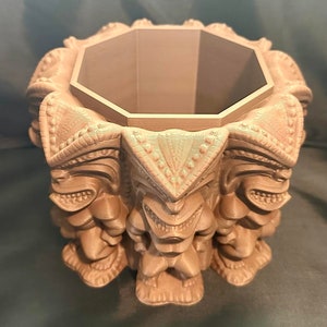 May include: A brown, 3D printed, octagonal planter with a tiki design. The planter is made up of eight tiki faces, each with a different expression. The planter is empty and ready to be filled with plants.