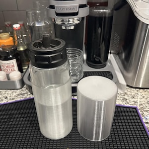 May include: A silver water bottle with a black lid and a silver cap. The bottle is sitting on a black and white checkered surface. There is a silver cap next to the bottle.