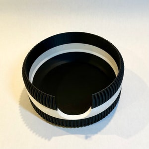 May include: Black and white round container with a ribbed edge and a white band around the top. The container has a cut-out on one side.
