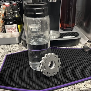May include: A clear glass bottle with a black lid filled with water sits on a black and purple mat. A silver gear-shaped object is in front of the bottle.