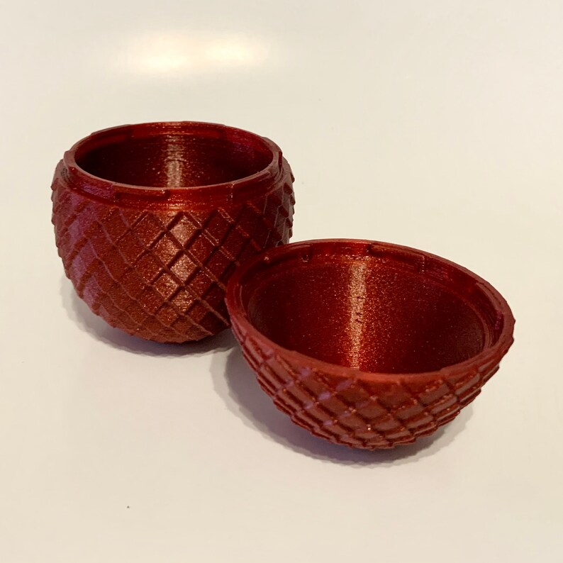 May include: A red, 3D printed, egg-shaped container with a textured, diamond-shaped pattern. The container is open and the two halves are separated.