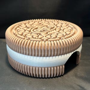 May include: A brown and white 3D printed Oreo cookie replica. The cookie has a detailed design and the text "OREO" is visible on the top.