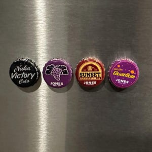 May include: Four colorful bottle caps with various designs and text. One cap reads "Nuka Victory Cola", another features a grape design, a third says "Sunset Sarsaparilla", and the last reads "Quantum Jones". All caps are from Jones Soda Co.