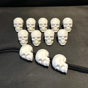 May include: White skull beads, some whole and some halved, are strung on a black cord. The beads are arranged on a dark gray surface. The skulls are a decorative element, possibly for jewelry or crafts.