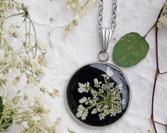 May include: A silver chain necklace with a round pendant featuring a pressed white flower encased in black resin.