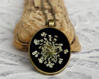 May include: A delicate white flower pressed inside a black oval pendant, set on a bronze chain. The pendant is on a piece of driftwood.