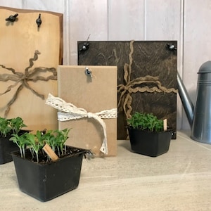 May include: Three wooden signs with twine and lace accents, two small black pots with green seedlings, and a silver watering can on a light brown tiled surface.
