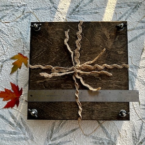 May include: A dark brown wooden box with a natural twine bow. The box is tied with a natural twine bow and has a metal ruler placed on top of it. The ruler is labeled "Westcott Flexible Stainless Steel Ruler" and has markings in inches.