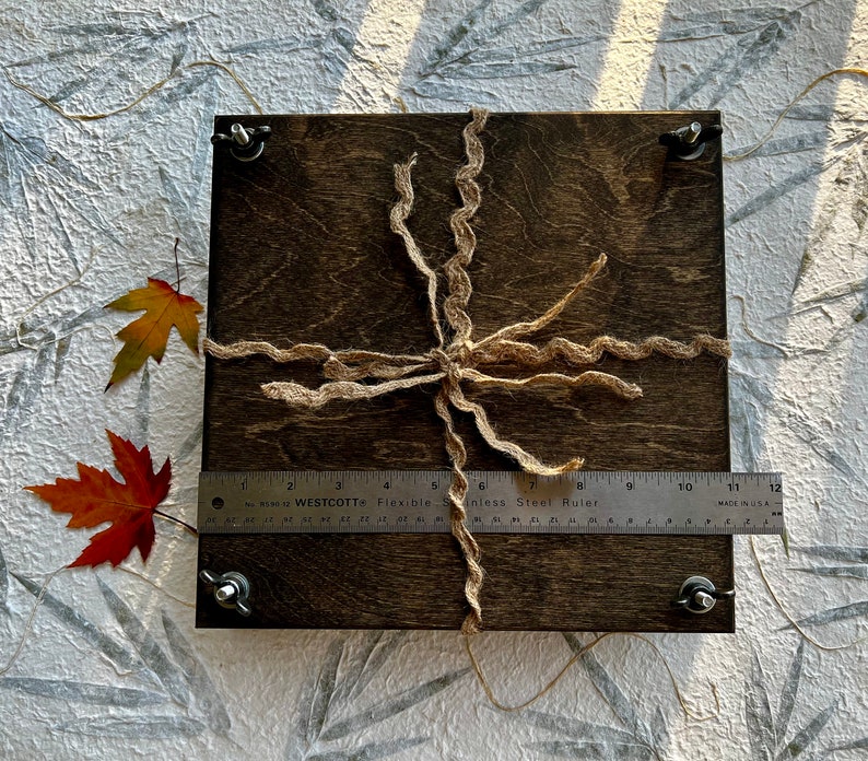 May include: A dark brown wooden box with a natural twine bow. The box is tied with a natural twine bow and has a metal ruler placed on top of it. The ruler is labeled "Westcott Flexible Stainless Steel Ruler" and has markings in inches.