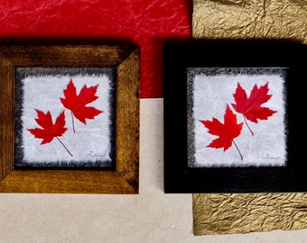 May include: Two framed red maple leaves on a white background. The frames are brown and black.