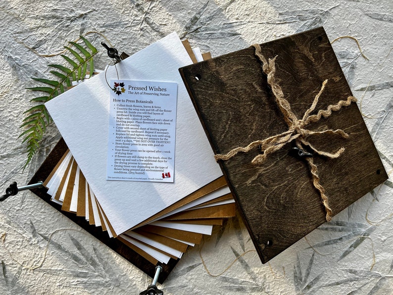 May include: A set of brown and white paper sheets for pressing flowers and botanicals. The sheets are stacked and held together with a metal clamp. The paper is packaged in a wooden box with a twine closure. The box has a label that reads "Pressed Wishes, The Art of Preserving Nature".