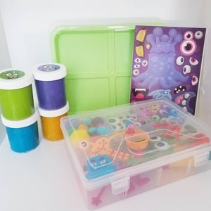 May include: A clear plastic storage container filled with colourful toy eyes, beads, and other small toys. There are three containers of play dough in the background, one green, one purple, and one orange. A sheet of paper with a monster design is also visible.