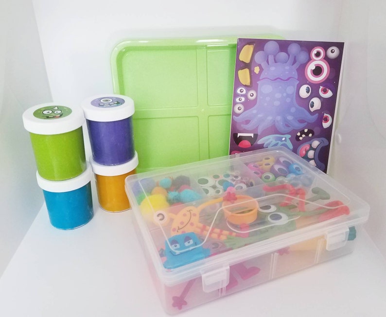 May include: A clear plastic storage container filled with colourful toy eyes, beads, and other small toys. There are three containers of play dough in the background, one green, one purple, and one orange. A sheet of paper with a monster design is also visible.