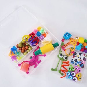 May include: Two clear plastic containers filled with colourful craft supplies. The first container has a rolling pin, pom poms, pipe cleaners, glitter, and a small monster toy. The second container has googly eyes, pom poms, and a green monster toy.