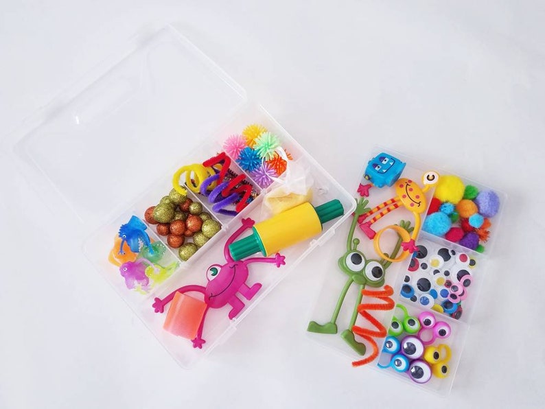 May include: Two clear plastic containers filled with colourful craft supplies. The first container has a rolling pin, pom poms, pipe cleaners, glitter, and a small monster toy. The second container has googly eyes, pom poms, and a green monster toy.