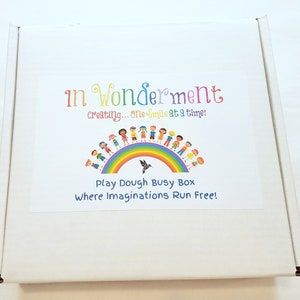 May include: A white cardboard box with a colourful label. The label features a rainbow with children holding hands around it. The text on the label reads "In Wonderment creating... one smile at a time! Play Dough Busy Box Where Imaginations Run Free!"