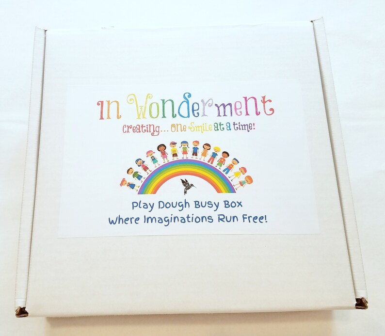 May include: A white cardboard box with a colourful label. The label features a rainbow with children holding hands around it. The text on the label reads "In Wonderment creating... one smile at a time! Play Dough Busy Box Where Imaginations Run Free!"