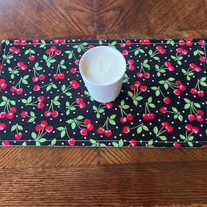 May include: A black and white fabric placemat with a cherry print and white polka dots. A white candle is sitting on the placemat.