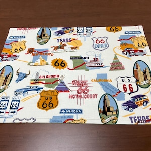 May include: Rectangular Route 66 themed placemat with a cream background. The design features illustrations of landmarks, vintage cars, and road signs. Text includes "Route 66", "Arizona", "Chicago", and "Texas".