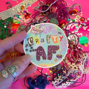 May include: A round sticker with a blue background and the words "Crafty AF" in colorful letters. The sticker is surrounded by glitter and other embellishments.