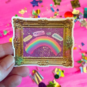 May include: A sticker with a rainbow and the words "dreams, magic, and glitter" in a gold frame. The background is purple with glitter.