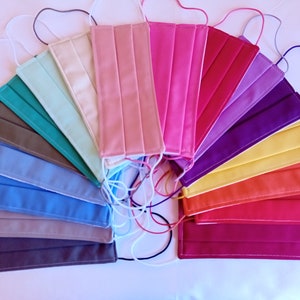 Op de afbeelding: Een verzameling rechthoekige stoffen gezichtsmaskers gerangschikt in een waaier. De maskers hebben verschillende effen kleuren, waaronder tinten roze, paars, geel, oranje, rood, blauw, groenblauw, grijs en beige, elk voorzien van elastische oorlussen.