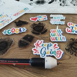K&ouml;nnte beinhalten: Eine Sammlung bunter Aufkleber mit motivierenden und inspirierenden Botschaften, darunter "Enjoy Today", "Think Positive", "Cool", "Okay Love", "Don't Waste Your Time", "Prove Them Wrong", "Well Well Well", "Keep Going" und "Highest Quality". Die Aufkleber befinden sich auf einer Holzoberfl&auml;che mit einem schwarzen und roten Werkzeug.