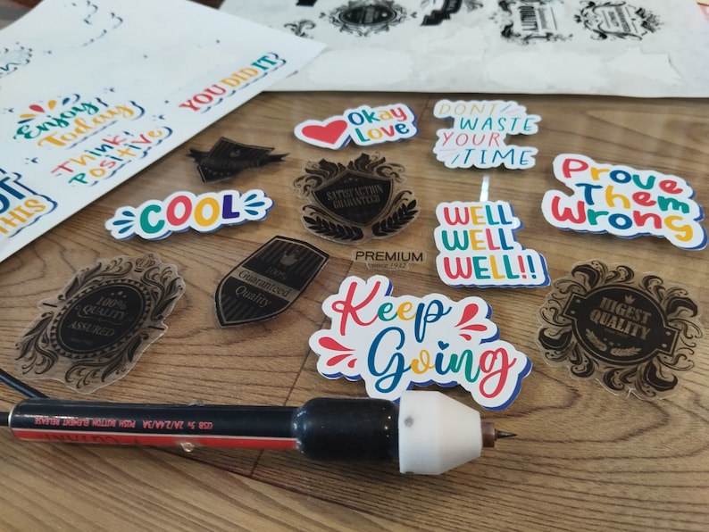 K&ouml;nnte beinhalten: Eine Sammlung bunter Aufkleber mit motivierenden und inspirierenden Botschaften, darunter "Enjoy Today", "Think Positive", "Cool", "Okay Love", "Don't Waste Your Time", "Prove Them Wrong", "Well Well Well", "Keep Going" und "Highest Quality". Die Aufkleber befinden sich auf einer Holzoberfl&auml;che mit einem schwarzen und roten Werkzeug.