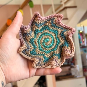 May include: A handmade, star-shaped crochet coaster. The coaster features a spiral design in shades of teal, tan, and gold, with a mauve border. The coaster is held in a hand, showcasing the intricate stitch work and color palette.