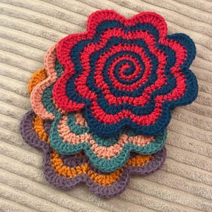 Puede incluir: Un conjunto de posavasos de flores de ganchillo hechos a mano en varios colores. El posavasos superior presenta un diseño en espiral rojo y azul marino. Otros posavasos incluyen turquesa, naranja, rosa y morado. Los posavasos están dispuestos sobre una superficie texturizada de color neutro.