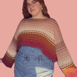 Puede incluir: Un suéter de crochet recortado y de tejido abierto en un degradado de colores, incluyendo tonos de rosa, marrón y naranja. El suéter tiene mangas largas y un escote amplio. La prenda se combina con un peto vaquero azul.