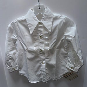 Pode incluir: Uma camisa branca de manga comprida com gola grande e frente com babados. A camisa tem fechos de botões na frente e nos punhos. A peça de roupa está pendurada num cabide branco.