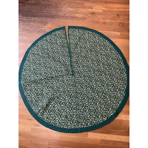 May include: A round Christmas tree skirt with a green and white floral pattern. The skirt has a dark green border and a slit down the center. The skirt is made of fabric and is designed to sit around the base of a Christmas tree.
