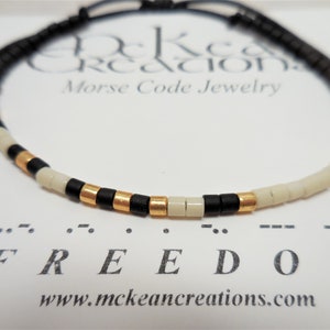 May include: A black cord bracelet with white and gold beads spelling out the word "FREEDOM" in Morse code. The bracelet is on a white card with the text "McKean Creations Morse Code Jewelry" and "www.mckeancreations.com".