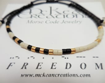 May include: A black cord bracelet with white and gold beads spelling out the word "FREEDOM" in Morse code. The bracelet is on a white card with the text "McKean Creations Morse Code Jewelry" and "www.mckeancreations.com".