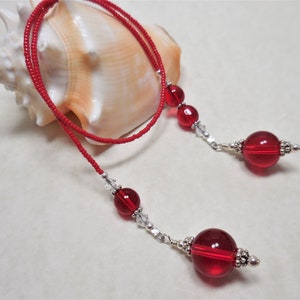 May include: A lariat necklace featuring a long strand of small red seed beads. The two ends are accented with larger translucent red glass spheres, smaller clear faceted crystal beads, and decorative silver-toned metal bead caps and spacers.
