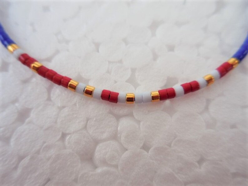 May include: A close-up of a red, white, and blue beaded necklace with gold accents. The beads are small and round, and the necklace is strung on a thin cord.