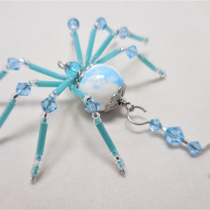 Puede incluir: Un adorno de ara&ntilde;a de cuentas turquesa y azul claro con un cuerpo de porcelana blanca y azul. La ara&ntilde;a tiene un colgador de alambre plateado con una gota de cuentas azul claro.