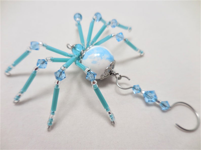 Puede incluir: Un adorno de ara&ntilde;a de cuentas turquesa y azul claro con un cuerpo de porcelana blanca y azul. La ara&ntilde;a tiene un colgador de alambre plateado con una gota de cuentas azul claro.