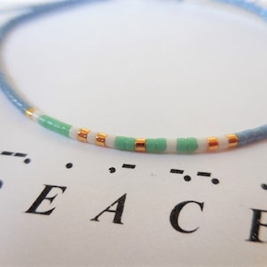 May include: A light blue bracelet with white and green beads and gold accents. The bracelet spells out the word "PEACE" in braille.