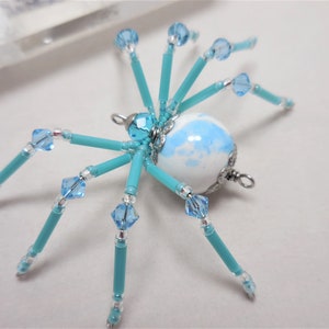 Puede incluir: Un adorno de ara&ntilde;a de cuentas turquesa con un cuerpo esf&eacute;rico de m&aacute;rmol blanco y azul y detalles de cristal.