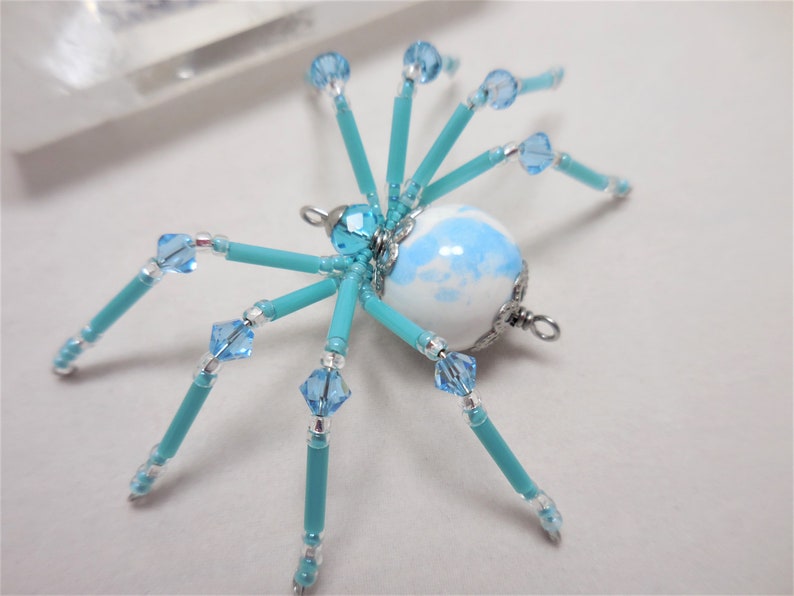 Puede incluir: Un adorno de ara&ntilde;a de cuentas turquesa con un cuerpo esf&eacute;rico de m&aacute;rmol blanco y azul y detalles de cristal.