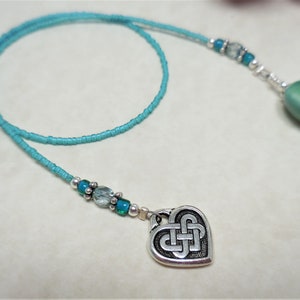 May include: A turquoise blue beaded bookmark with a silver heart charm featuring a Celtic knot design. The bookmark also has a green ceramic bead at the end.