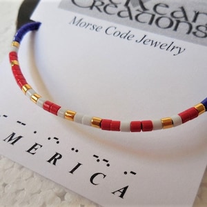 May include: A red, white, and blue beaded bracelet with gold accents. The bracelet spells out "AMERICA" in Morse code.