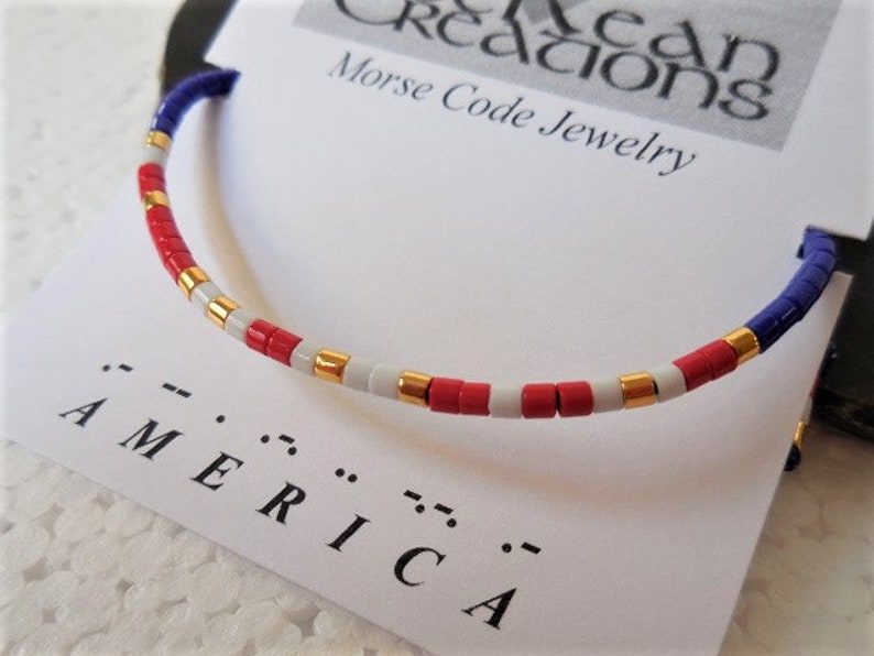 May include: A red, white, and blue beaded bracelet with gold accents. The bracelet spells out "AMERICA" in Morse code.