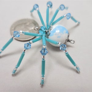 Puede incluir: Un adorno de ara&ntilde;a de cuentas con un cuerpo estampado en blanco y azul, patas turquesa y detalles plateados. La ara&ntilde;a es del tama&ntilde;o aproximado de una moneda de 25 centavos de d&oacute;lar estadounidense.