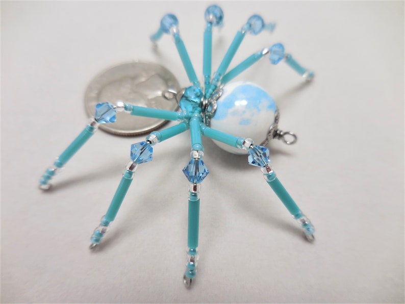 Puede incluir: Un adorno de ara&ntilde;a de cuentas con un cuerpo estampado en blanco y azul, patas turquesa y detalles plateados. La ara&ntilde;a es del tama&ntilde;o aproximado de una moneda de 25 centavos de d&oacute;lar estadounidense.