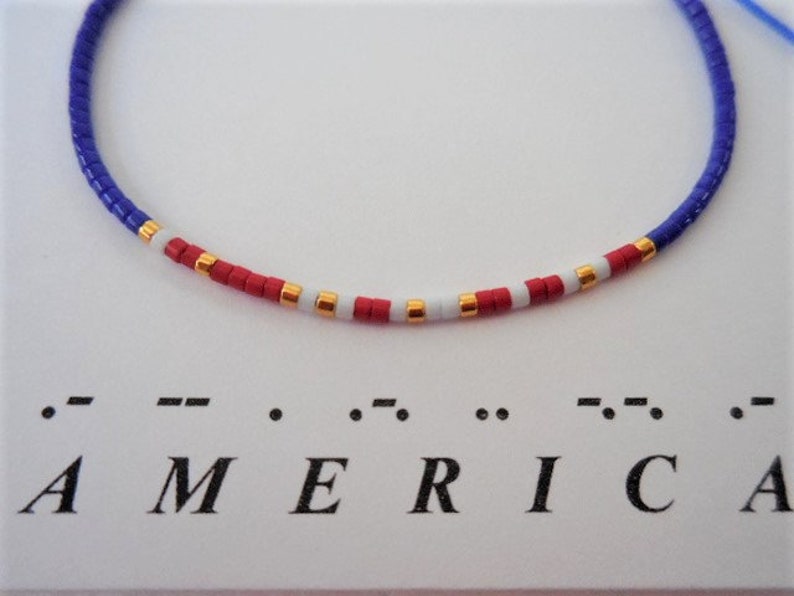 May include: A blue, red, white, and gold beaded bracelet with the word "AMERICA" spelled out in braille below it.
