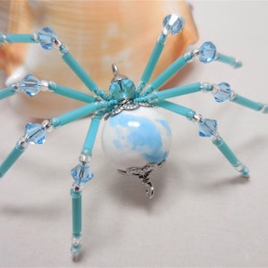 Puede incluir: Un amuleto de ara&ntilde;a de cuentas con un cuerpo esf&eacute;rico de m&aacute;rmol azul y blanco, patas turquesa y detalles de cristal transparente.
